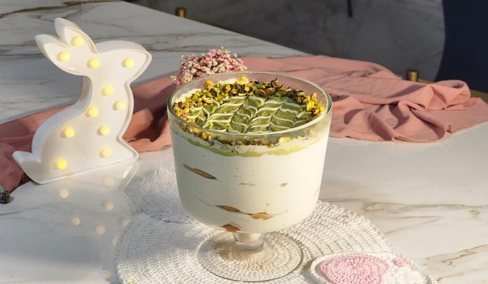 Fotografia de uma taça de vidro servido com camadas de creme, biscoito e pasta de pistache. Ao fundo, um pano rosa e uma luminária em formato de coelho.