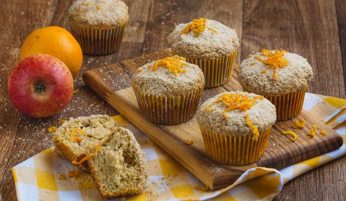 Imagem aproximada de muffins em tom claro, sobre uma tábua de madeira em um pano quadriculado em tom amarelo e branco. Ao fundo há uma maçã e uma laranja e alguns farelos de Farinha Láctea.