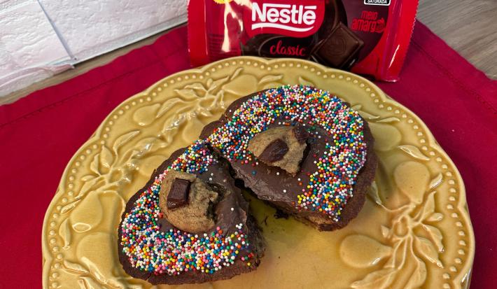 Fotografia em tons de vermelho com um prato amarelo ao centro. Em cima do prato existe uma receita de ovo de páscoa feita com a casca de cookie e recheado com brigadeiro. Na finalização, existe cookies e granulados coloridos