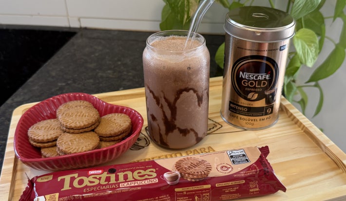 Foto da receita de Frappuccino de Chocolate com Biscoito Tostines, servido em um copo com canudo, sobre uma bandeja de madeira com Biscoitos Tostines Cappuccino soltos e o pacote à frente e, do lado direito, uma lata de Nescafé Gold