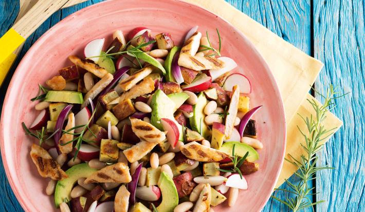 Fotografia em tons de azul e rosa em uma bancada de madeira azul com um prato rosa com salada de batata-doce, frango, feijão branco e abacate dentro dele.