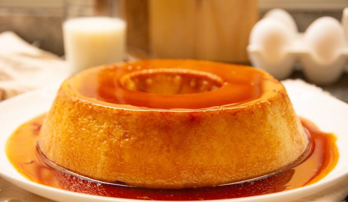 Fotografia mostra uma torta com textura delicada e cremosa, coberta por uma calda de caramelo dourada e servida em um prato branco.