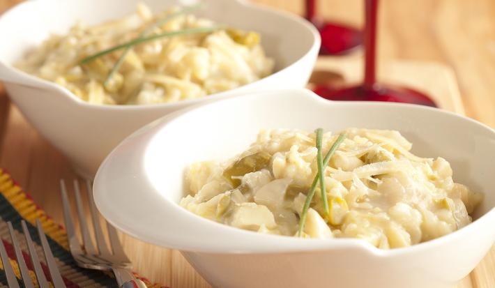 foto em tons de marrom, branco e vermelho tirada de uma bancada vista de frente, contém dois recipientes redondos brancos com o risoto com aspargos. Ao fundo duas taças vermelhas e à frente dois garfos para servir.