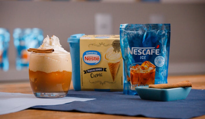 Copo com bebida cremosa ao lado de um sachê azul de NESCAFÉ® Ice e um prato com canela em pau.