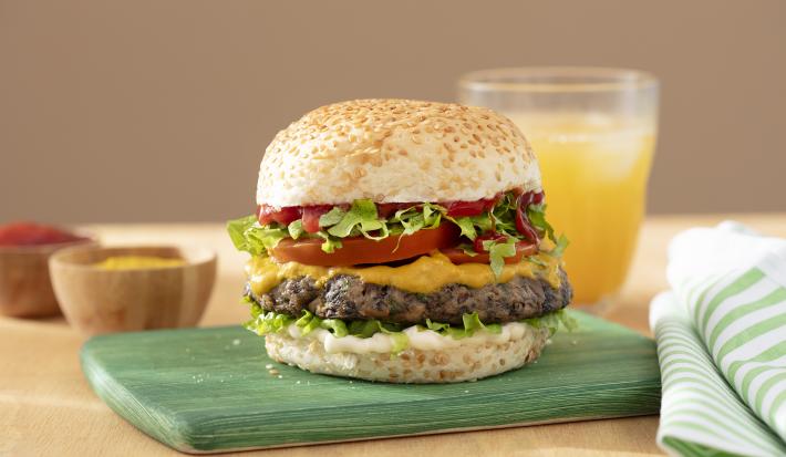 Foto da receita de Hambúrguer de Cogumelo. Observa-se um lanche montado com pão de hambúrguer com gergelim, mostarda, ketchup, alface, tomate e o hambúrguer de cogumelo ao meio.