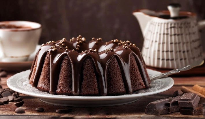 Em cima de uma mesa, prato redondo com bolo de chocolate com furo no meio e cobertura de chocolate cremoso.