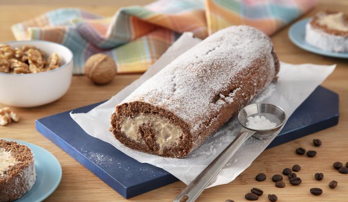 Foto da Receita de Rocambole de Chocolate com Café. Observa-se o rocambola sobre uma tábua azul escura e uma superfície de madeira. Grãos de café decoram a foto, assim como nozes
