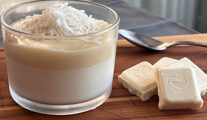 Imagem mostra um copo pequeno transparente com um creme branco por baixo e um creme bege por cima com coco ralado. Uma colher prateada está ao lado, junto de quadradinhos de chocolate branco. Tudo está apoiado em uma tábua de madeira.