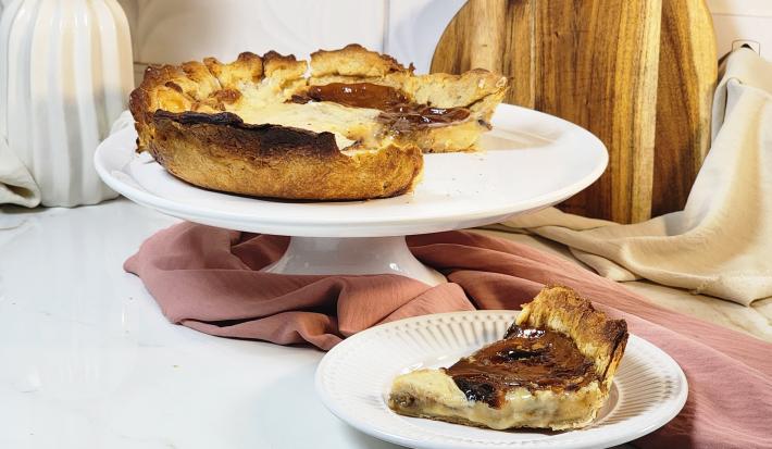Fotografia em tons de marrom em uma bancada de mármore com um pano marrom em volta e um prato branco elevado com a torta fácil de café ao centro e ao lado um pratinho com um pedaço  da torta. Ao fundo, uma tábua de madeira.