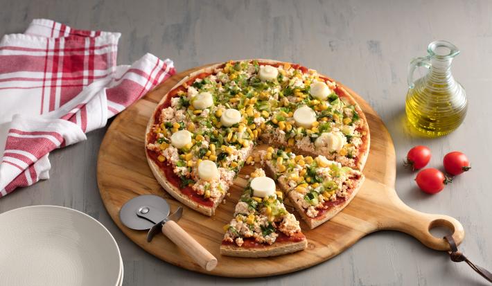 Fotografia em tons de vermelho em uma bancada de madeira cinza com uma tábua de pizza de madeira com a pizza em cima decorada com escarola e ricota. Ao lado, um vidro de azeite, tomates e um cortador de pizza.