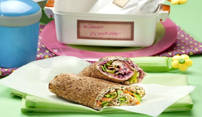 Fotografia em tons de verde em uma mesa verde com um pano verde, um papel manteiga com dois wraps recheados com salada e cebola. Ao fundo, uma marmita branca com detalhes rosa.