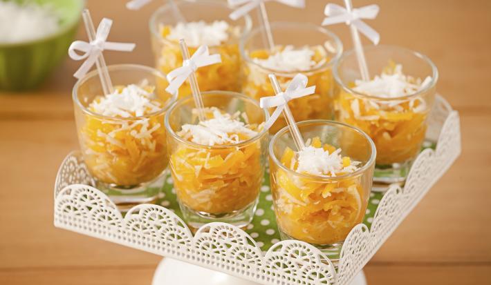 Fotografia em tons de laranja em uma mesa de madeira com um suporte de doces branco ao centro com os doces de abóbora em copinhos transparentes com palitinhos e um lacinho branco.