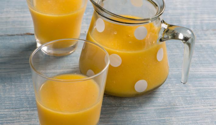 Fotografia em tons de azul e amarelo com uma jarra de vidro com bolinhas brancas e dois copos contendo suco de frutas, tudo sobre bancada em tons de azul.