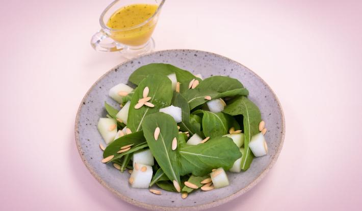 Fotografia em tons de rosa e verde em uma mesa de madeira rosa com um prato redondo raso lilás e a salada de rúcula com limão dentro dele. Ao fundo, um potinho com azeite.