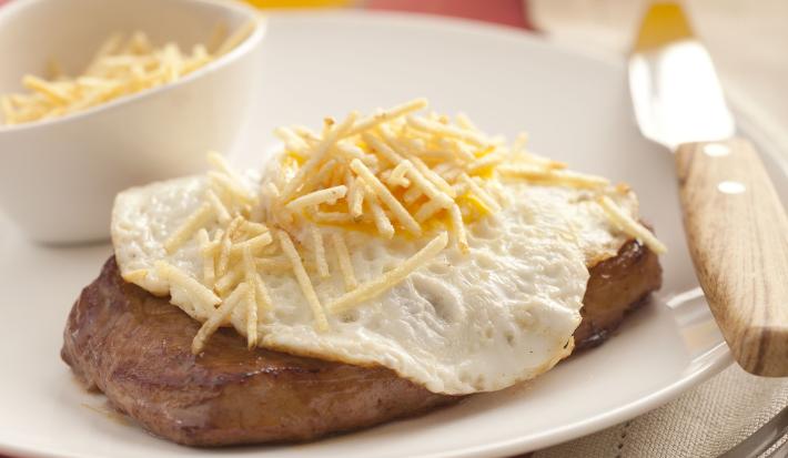 fotografia em tons de rosa, branco e marrom de uma bancada rosa vista de frente, contém um prato redondo branco com um pedaço de carne com ovo por cima e batata palha. Ao lado garfo e faca para servir, e ao fundo uma jarra e um copo transparente com suco.