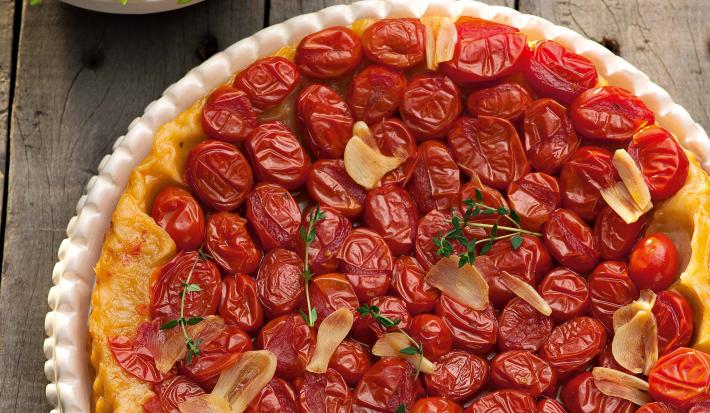 fotografia em tons de cinza, vermelho e verde de uma bancada cinza vista de cima. Contém um recipiente branco redondo com a torta e diversos tomates-cereja por cima