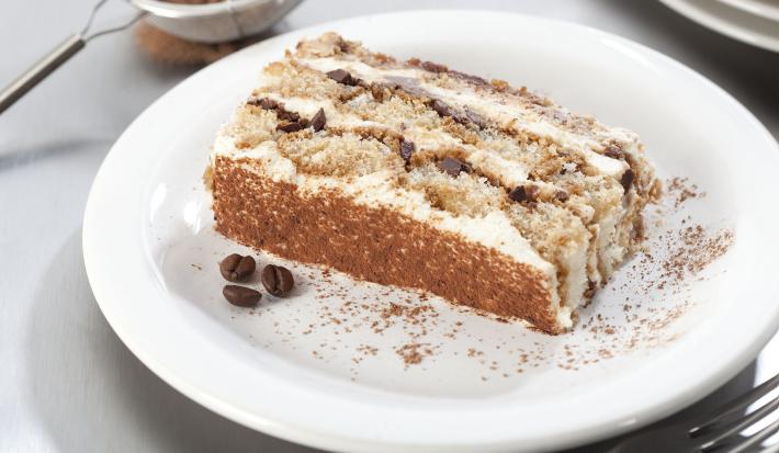 Fotografia em tons de branco, marrom e cinza, com prato branco ao centro com porção de bolo fatiado, no entorno garfo, peneirinha com chocolate em pó e pratos brancos empilhados, tudo sobre bancada em tons de cinza.