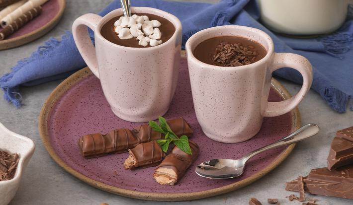 Foto de duas canecas em tom rosa claro, contendo a receita de Chocolate Quente, sobre um prato em tom violeta, com um tecido em tom azul atrás, uma jarra com leite e pedaços de chocolate e canelas em pau sobre a mesa