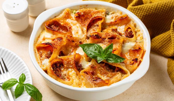 Fotografia de um recipiente branco de caçarola com uma receita de conchiglione gratinado. O recipiente está sobre uma mesa de cor clara, ao lado de um pano de tom mostarda e um prato branco raso com um garfo.