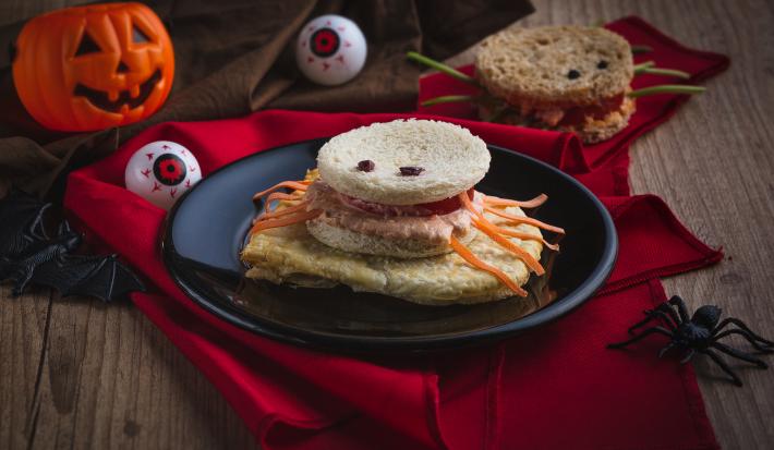 Imagem aproximada da receita de um sanduíche em formato de aranha, com pão, patas feitas com tiras de cenoura, além de o cenário estar decorado com itens de Halloween