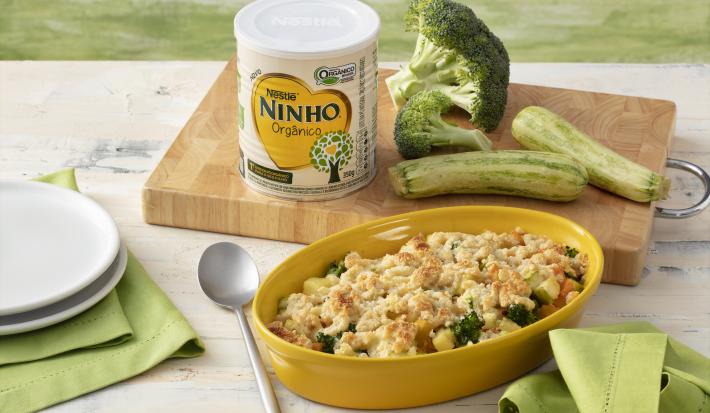 Fotografia em tons de verde e branco de uma bancada branca sobre ela um recipiente oval amarelo com o crumble de legumes. Ao lado um paninho verde, dois pratos brancos e uma colher. Ao fundo uma tábua de madeira com uma lata de NINHO, brócolis e abobrinha