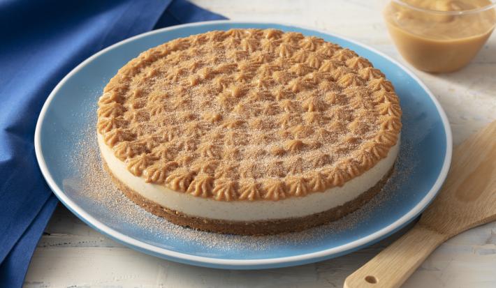 Foto de uma bancada clara. Sobre ela há uma prato azul com a receita de Torta pronta, feita com doce de leite e polvilhada com açúcar e canela; há uma espátula de madeira, um tecido azul e um pote com doce de leite também compondo a cena
