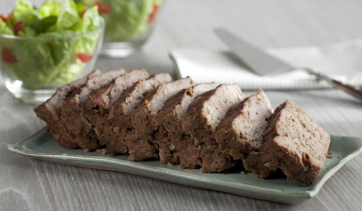 Fotografia em tons de marrom e cinza, com bolo de carne assada sobre peça de porcelana cinza e no entorno guardanapo branco com espátula por cima e dois potinhos com folhas verdes, tudo sobre bancada em tons de cinza.