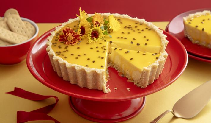 Foto da receita de Cheesecake de Maracujá e Chocolate Branco. Observa-se uma torta com calda de maracujá e flores comestíveis com um pedaço cortado, em cima de uma boleira vermelha.