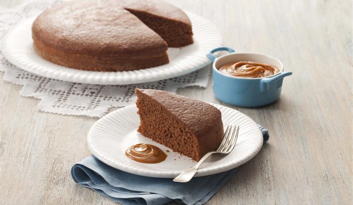 fotografia em tons de cinza e marrom de uma bancada cinza vista de frente. Contém um prato redondo branco, com um bolo de chocolate sem uma fatia. Ao lado outro prato com a fatia de bolo e um garfo ao lado para servir.
