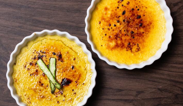 Fotografia vista de cima de dois ramekins brancos, um com creme brulée de baunilha com laranja, e o outro creme brulée de aspargos com bacon. Os ramekins estão sobre uma mesa de madeira.