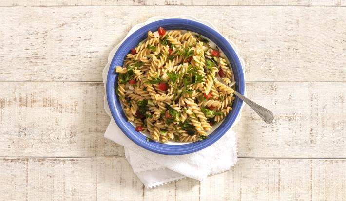 em uma mesa de madeira na cor cinza, contém um recipiente na cor azul que comporta a salada de macarrão.