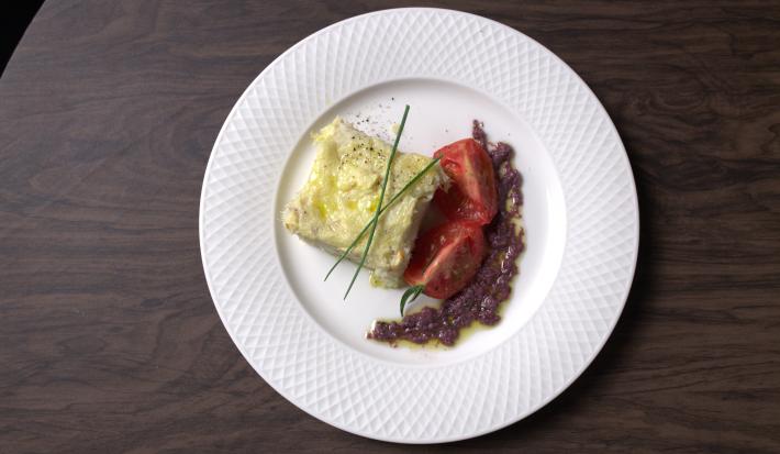 Fotografia em tons de branco com um prato branco ao centro. Dentro do prato existe uma receita de bacalhau acompanhada por tomates e azeitonas