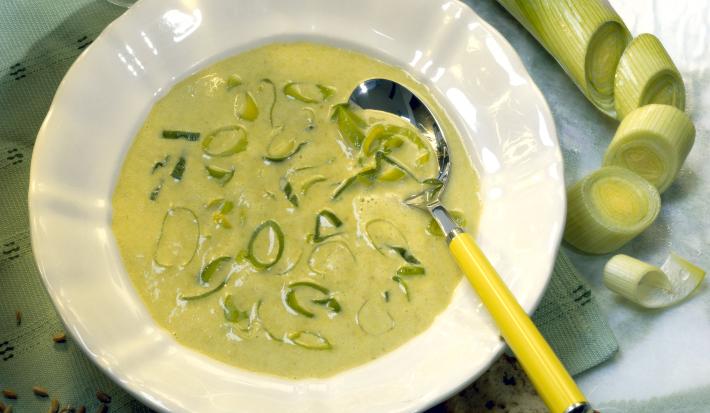 Fotografia de um prato branco fundo com um caldo de alho poró, com uma colher de cabo amarelo apoiada sobre a sopa, que tem pedaços de alho poró em cima. Ao redor, um alho poró inteiro, duas fatias de pão, uma taça vazia e grãos, sobre uma mesa verde.