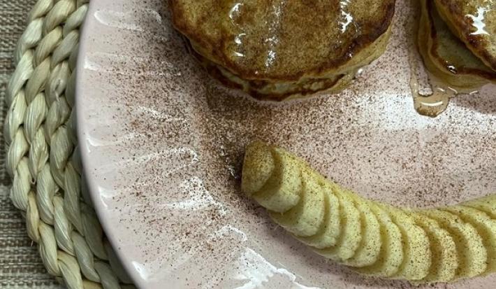 Foto da receita de Panquequinha de Aveia. Observa-se um prato com panquequinhas com mel e canela e do lado de baixo do prato, uma banana em rodelas para formar um sorriso.