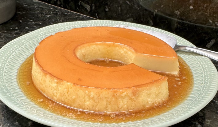 Imagem de um pudim lindo, com uma fatia faltando, sobre um prato verde claro e com a aparência de mais furadinho