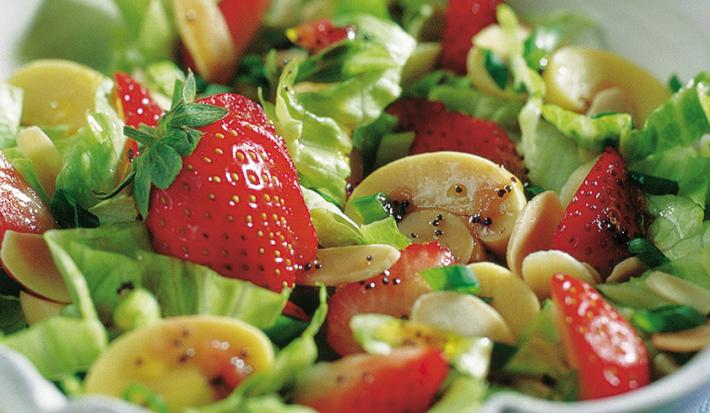Fotografia em tons de verde em uma bancada de madeira azul com um recipiente redondo e fundo com a salada primavera composta por alface, cogumelo e morango.