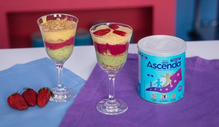 Fotografia em tons claros de duas taças de vidro com os cremes coloridos intercalados com o biscoito nesfit. As taças estão sobre dois panis, azul e roxo, ao lado tem a lata de ascenda e três morangos inteiros.