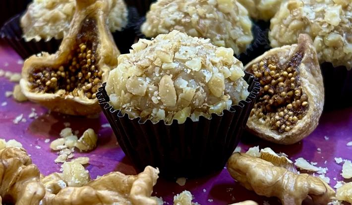 Foto de receita de Brigadeiro de Figo com Nozes. Observa-se o brigadeiro com nozes picadas em volta em uma forminha preta, com figos secos cortados ao lado e nozes na projeção da frente. Ao fundo, mais brigadeiros.