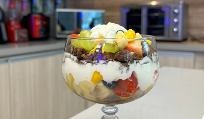 Fotografia de uma taça grande de vidro com pavê de frutas e iogurte, O pavê está sobre uma bancada e no fundo tem aparelhos eletrodomésticos.
