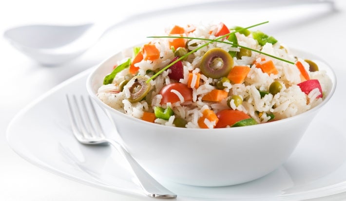 Em cima de uma mesa, tigela redonda com porção de arroz soltinho, tomate picado, cenoura, ervilha e azeitonas verdes.