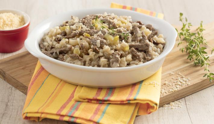 Fotografia de um recipiente redondo com alças brancas e um guardanapo amarelo com listras coloridas, dentro dele pedaços o risoto com carne e folhas verdes decorativas. Ao fundo um potinho vermelho com queijo ralado.