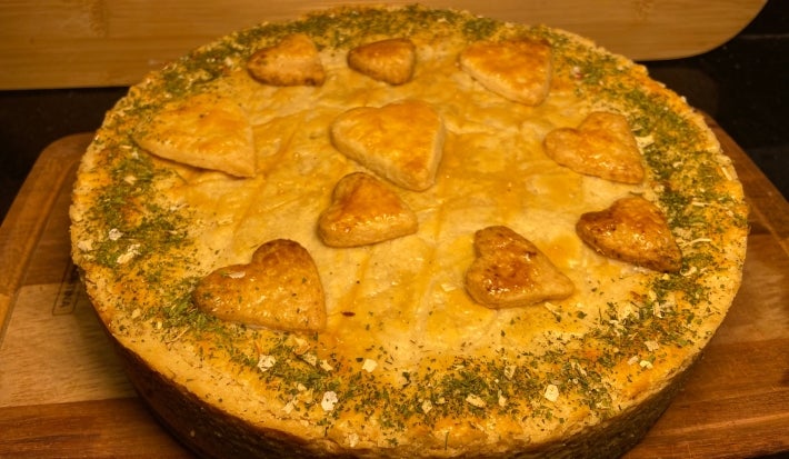 Fotografia com luzes mais amareladas e baixas de um empadão inteiro recheado com franog e requeijão sobre uma tábua retangular. O empadão tem alguns coraçãoes de massa para decorar e tem algumas ervas finas salpicando as bordas da torta.
