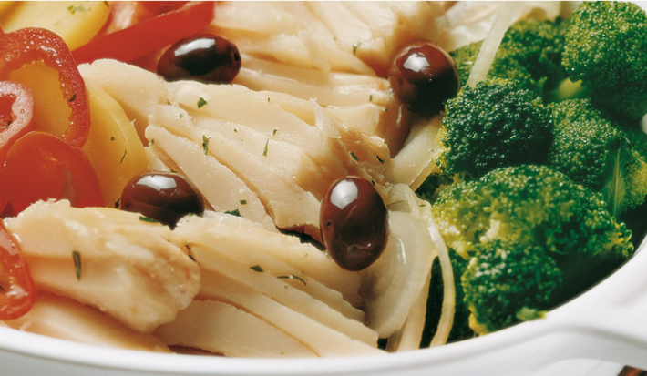 Fotografia em tons de verde e vermelho em uma bancada de madeira em uma mesa com uma toalha cinza, um refratário oval branco com bacalhau com tomate, batata e brócolis.
