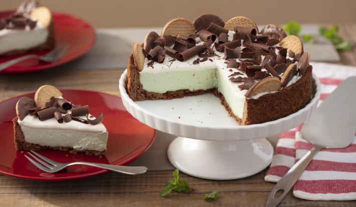 Foto da receita de Torta de Calipso e Menta, em tons marrom claro e verde claro, sobre um prato branco alto, numa bancada de madeira onde há mais um prato vermelho com uma fatia da torta, um tecido vermelho e branco e uma espátula de bolos