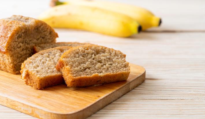 Imagem de fatias de um bolo retangular de banana sobre uma tábua de madeira e ao fundo duas bananas.