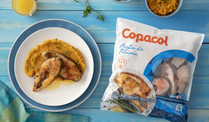 Foto da receita de Posta de Tilápia Copacol com Molho Cítrico e Purê de Abóbora, vista de cima, servida em um prato redondo, sobre uma bancada azul decorada com ingredientes e um tecido, ao lado do produto Copacol