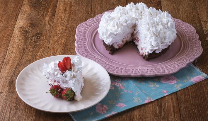 Imagem da receita de Torta Brownie, decorada com chantilly e morangos, servida em um prato rosa e ao lado há um prato menor branco com uma fatia. Tudo está sobre um pano decorado com motivos florais, sobre uma bancada de madeira