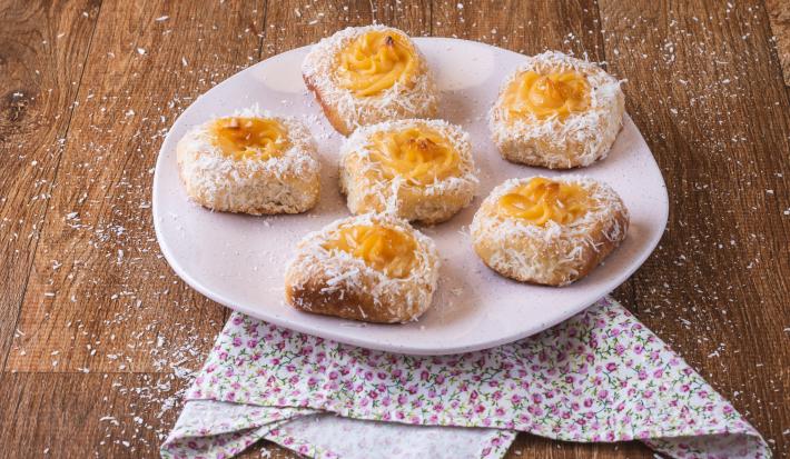 Foto da receita de pão com creme de leite moça, servidos em um prato rosa claro, com coco ralado salpicado, em uma bancada de madeira decorada com um pano de motivos florais