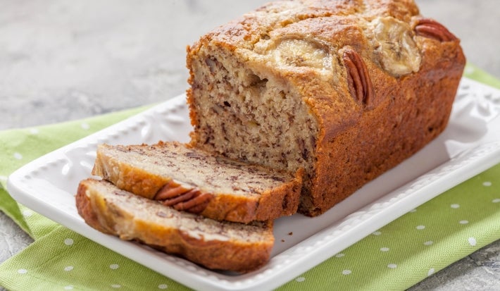 Fotografia em tons claros de um bolo de banana integral feito em forma de bolo inglês, com bananas e castanha decorando o bolo, que está sobre um apoio retangular branco, com duas fatias cortadas.