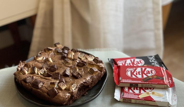 Foto aproximada de um Brownie de KitKat, em formato quadrado, sobre um prato preto. Ao lado dele há três KitKtas, um ao leite, outro dark e outro white.